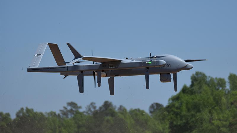Aerosonde flying in the sky with the tree line in the background. 
